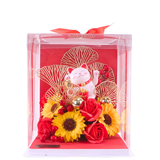 A packaged artificial flower arrangement featuring red and yellow flowers with a white cat figurine, presented in a clear box with a red ribbon.